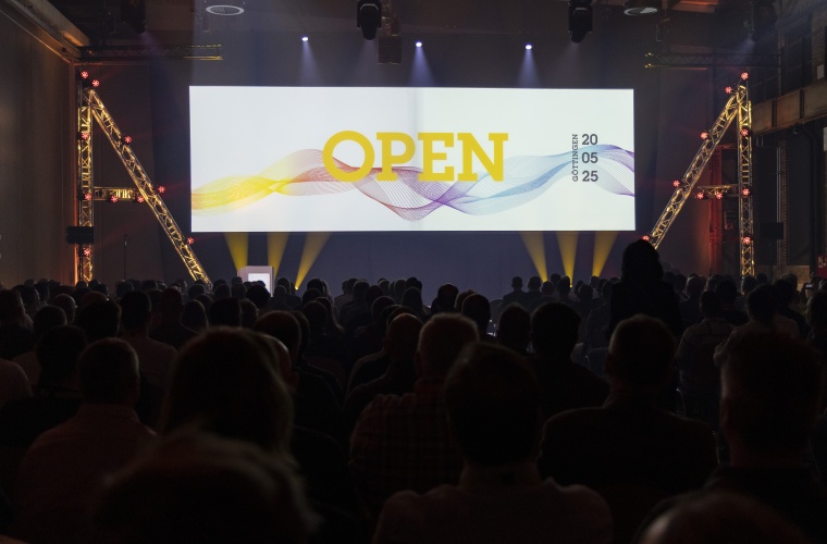 Blick von hinten auf einen vollen und abgedunkelten Plenarsaal. Auf der Bühne ist die Aufschrift „OPEN“ zu lesen