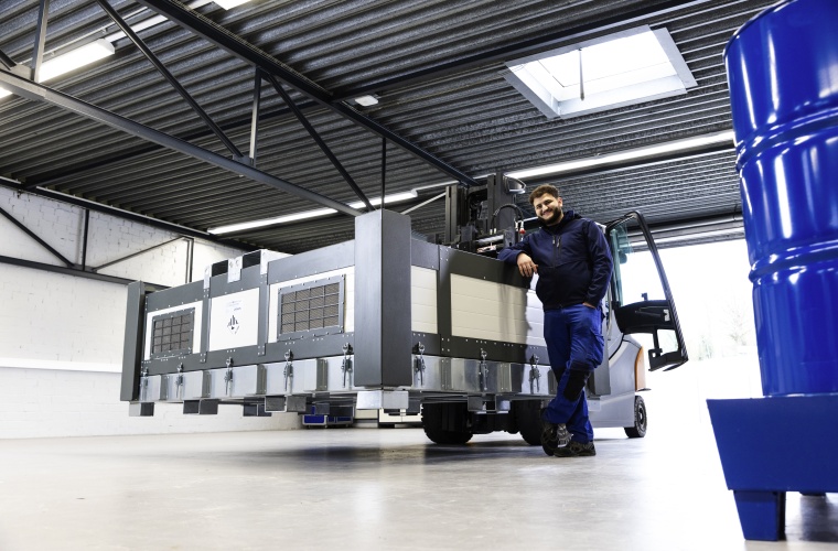 Person lehnt an großer Industriemaschine in einer Halle; im Vordergrund ein blauer Fassbehälter, Decke mit Stahlträgern