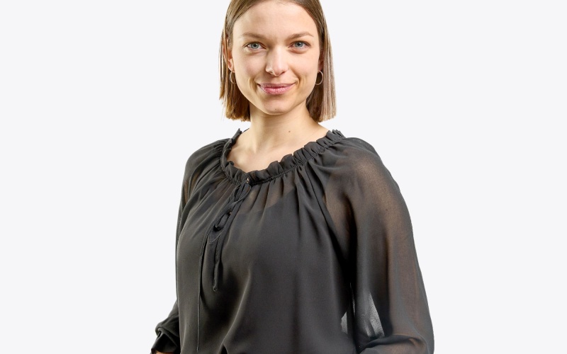 Eine Person steht vor einem neutralen Hintergrund und trägt eine schwarze, locker fallende Bluse mit gerafftem Ausschnitt und weiten Ärmeln sowie eine blaue Jeans mit schwarzem Gürtel. Die Hände sind in die Hosentaschen gesteckt