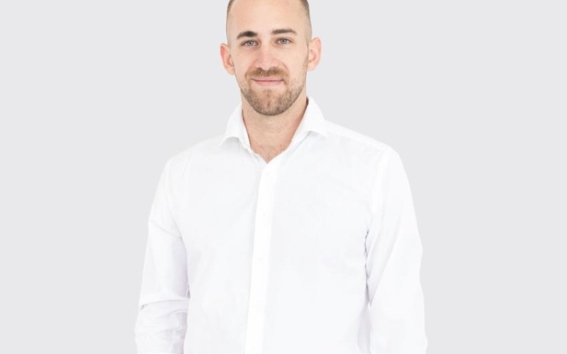 Eine Person steht vor einem neutralen, hellgrauen Hintergrund und trägt ein weißes, langärmliges Hemd mit hochgekrempelten Ärmeln sowie eine beige Hose mit schwarzem Gürtel