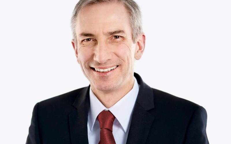 Eine Person ist vor einem hellen Hintergrund abgebildet und trägt einen dunklen Anzug mit weißem Hemd und einer roten Krawatte