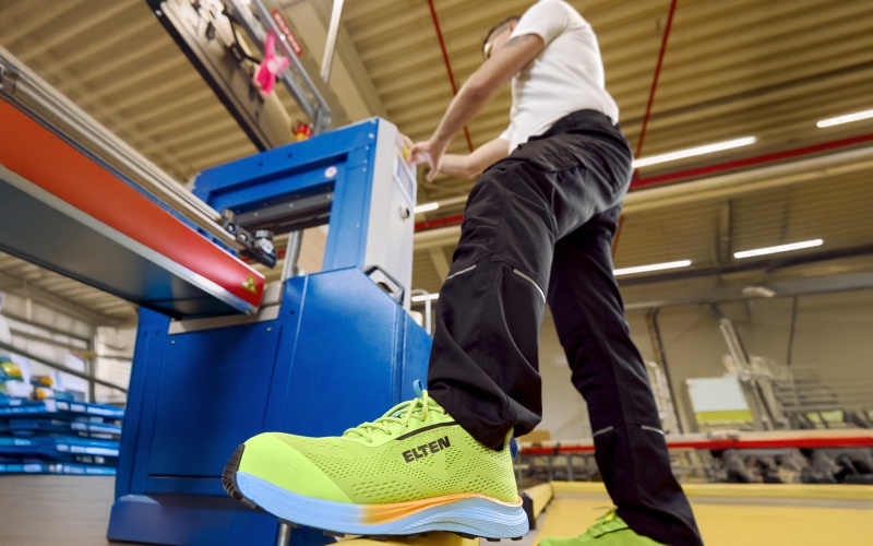 Person arbeitet an einer Maschine; im Vordergrund ein neongrüner Sicherheitsschuh