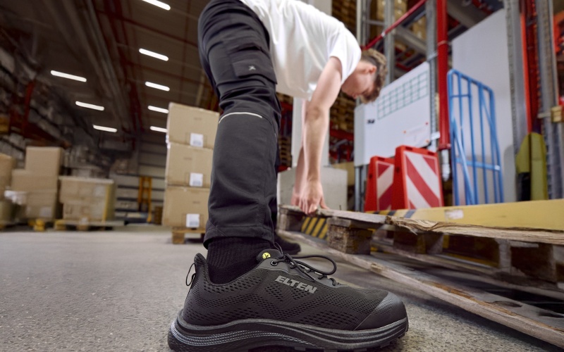 Person bewegt eine Palette in einer Lagerhalle; im Vordergrund ein schwarzer Sicherheitsschuh
