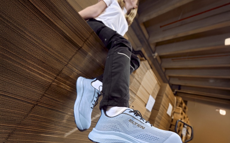 Person sitzt auf gestapeltem Material in einer Halle; im Vordergrund ein hellblauer Sicherheitsschuh