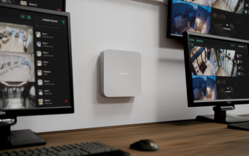 Quadratisches, weißes Wandgerät mit abgerundeten Ecken mit der Aufschrift Ajax – mit diversen Monitoren daneben