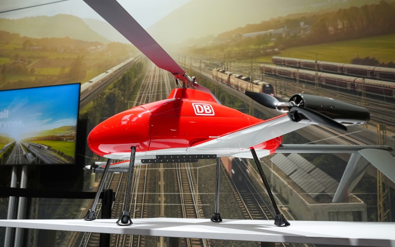 Rotglänzende Drohne mit zwei großen Rotorblättern, Aufschrift DB, vor Bild mit Bahngleisen