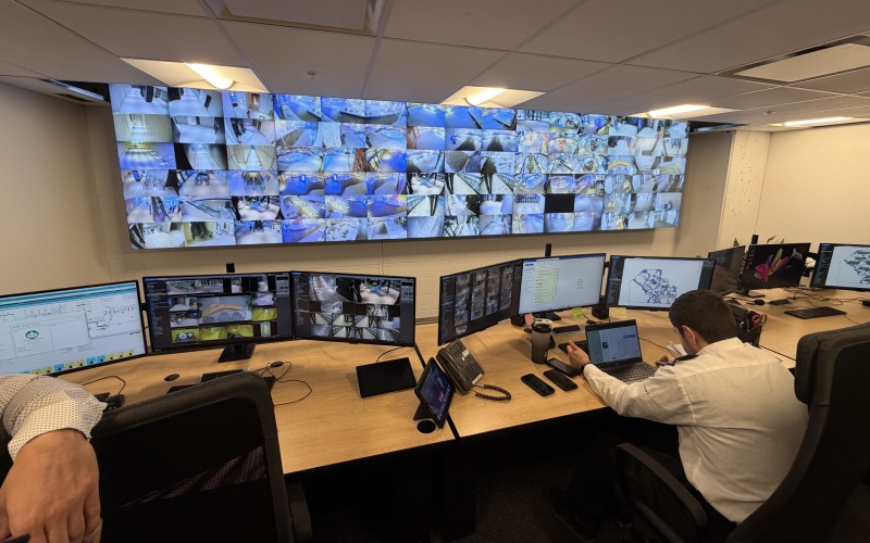 Mann der vor mehreren Monitoren sitzt, dahinter eine Wand mit Splitbildschirmen