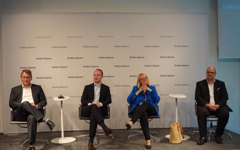 Podiumsdiskussion mit vier sitzenden Personen auf einer Bühne vor einer gebrandeten Veranstaltungswand und kleinen Stehtischen