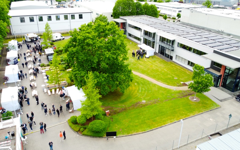 Drohnenaufnahme eines Firmengeländes mit modernem Bürogebäude, Grünflächen und einem Außenbereich, in dem viele Personen zwischen weißen Pavillons unterwegs sind
