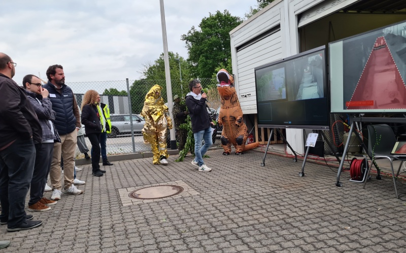 Alternativtext: Mehrere Personen stehen im Freien auf einem Betriebsgelände und betrachten zwei Monitore vor einem geöffneten Anhänger; einige tragen auffällige Schutz- und Kostümanzüge, darunter ein goldfarbener Hitzeschutzanzug und ein aufblasbares Dinosaurierkostüm