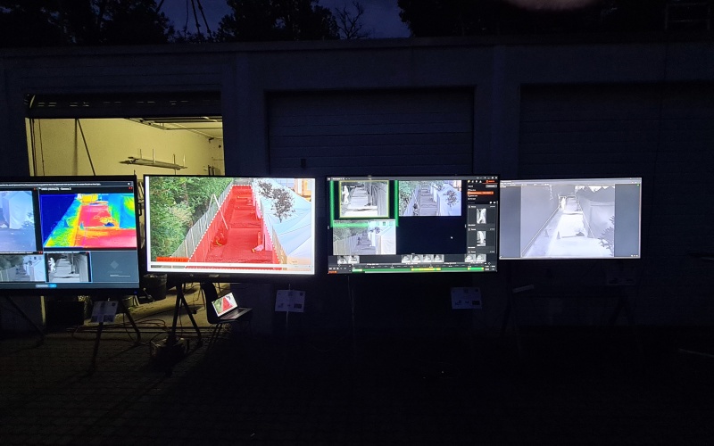 Mehrere Monitore stehen nachts im Freien vor einer Halle und zeigen verschiedene Kameraansichten, darunter Wärmebild-, Farb- und Schwarzweißaufnahmen eines abgesperrten Testbereichs