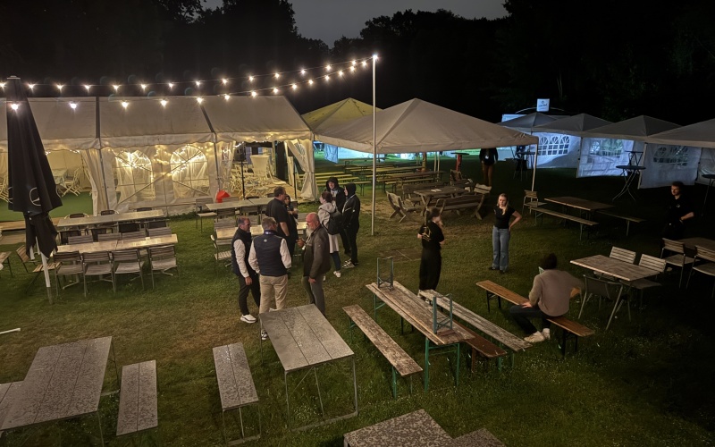 Sommerfest bei Nacht mit mehreren beleuchteten Zelten, langen Biertischen und einigen stehenden und sitzenden Personen auf einer Wiese