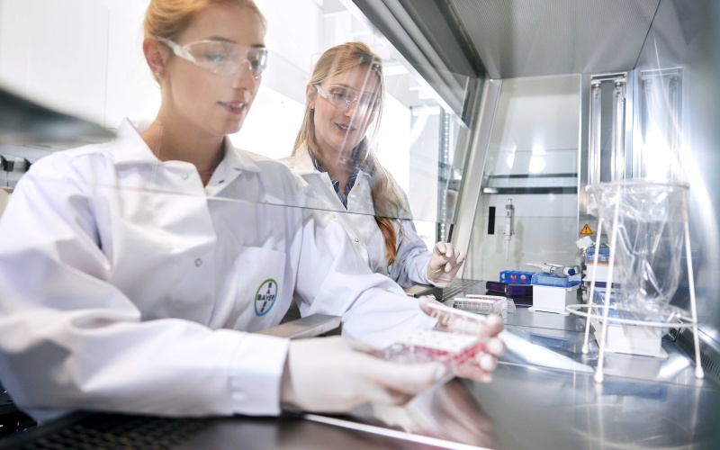Zwei Mitarbeiterinnen in weißen Kitteln vor der Glasschutzscheibe eines Chemie-Arbeitsplatzes
