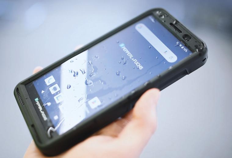 Hand hält ein schwarzes Smartphone mit Wassertropfen auf dem Display; auf dem...