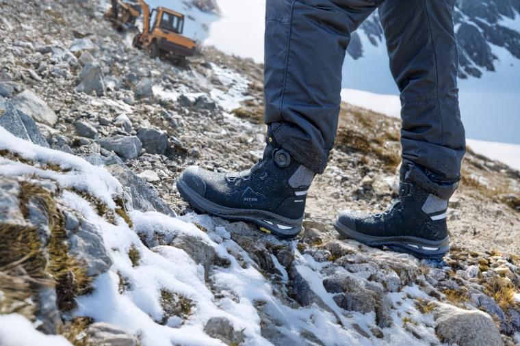 Eine Person in blauen Wanderstiefeln geht einen schneebedeckten, felsigen Hang...