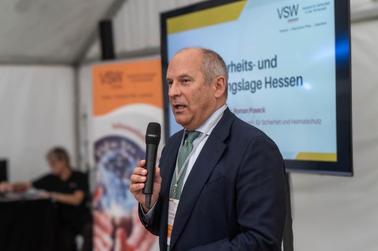 Ein Mann im Anzug spricht mit Mikrofon vor einem Bildschirm mit der Aufschrift...