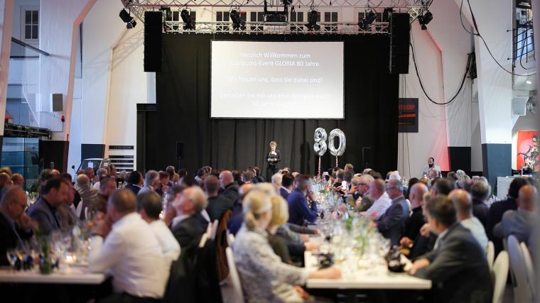 Ein großer Saal mit Bühne, einem Ballon in Form einer 80 sowie Gästen an...