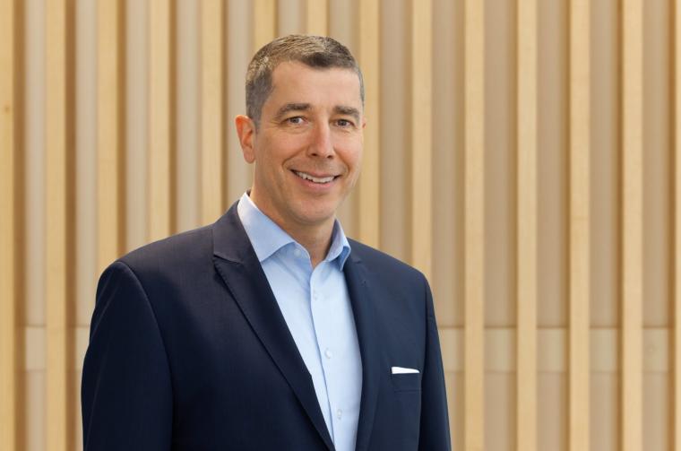 Portrait eines mittelalten Mannes im blauen Sakko vor hölzerner Lamellenwand