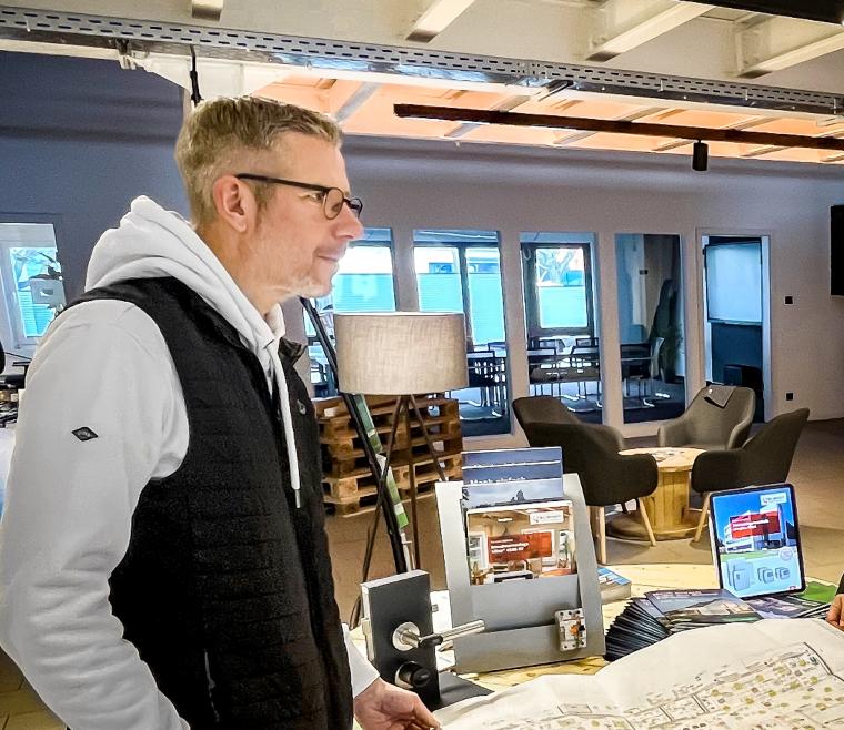 Mann mit Brille und Hoody mit Planungs- und Werbematerial, dahinter Sitzgruppe...