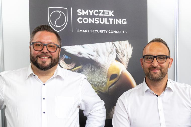 Zwei Männer mit weißen Hemden vor einem Plakat mit einem Falken und der...