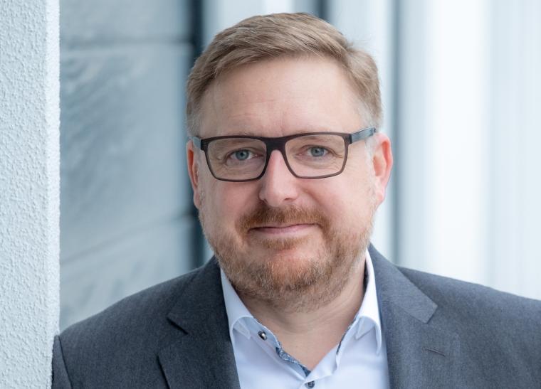 Person in grauem Sakko und weißem Hemd lehnt an einer hellen Außenwand