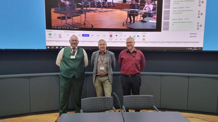 Drei Herren vor großer Projektion einer Präsentation mit Screenshot