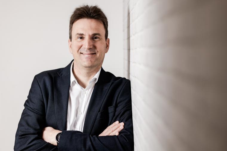 Person steht mit verschränkten Armen an einer hellen Wand in einem Innenraum.