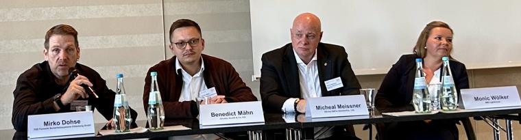 Drei mittelalte Männer und eine junge Frau an einem Podiumstisch, alle in eine...