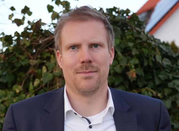 Person in Sakko und Hemd steht im Freien vor einer Hecke