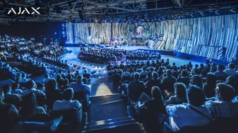 Large auditorium with a stage lit in blue, a circular backdrop behind speakers,...
