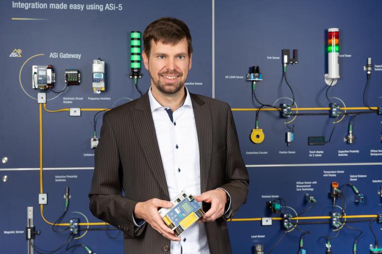Person in Sakko hält ein elektronisches Modul vor einer Wand mit vernetzter...