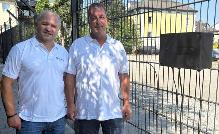 Zwei Personen in weißen Firmenpoloshirts stehen neben einem gesicherten Zaun...