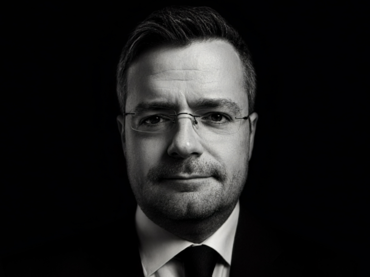 Mann mit randloser Brille, schwarzer Anzugjacke und schwarzer Krawatte