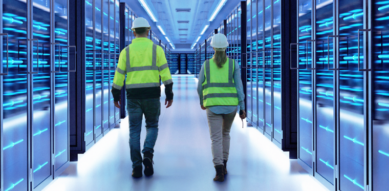 Two people wearing protective vests walk between blue-lit racks in a data center