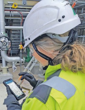 Eine Person mit weißem Schutzhelm und gelber Warnschutzjacke bedient ein Smartphone auf einem Industriegelände. Im Hintergrund sind Rohre, Ventile und Messgeräte zu sehen