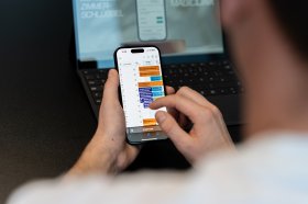 Aufnahme über die Schulter eines Manns der sein Handy bedient. Im Hintergrund ist ein aufgeklappter, eingeschalteter Laptop zu sehen