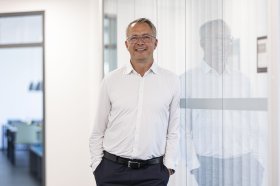 Ein Mann mit kurzen grauen Haaren, Brille, weißem Hemd, dunkelblauer Hose mit schwarzem Gürtel, der sich gegen eine Raumverglasung mit der linken Schulter lehnt
