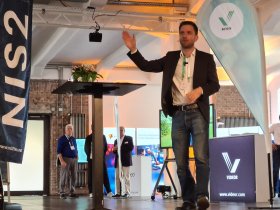 Eine Person steht auf einer Bühne und hält eine Präsentation. Sie trägt einen dunklen Blazer, ein weißes Hemd und Jeans. Im Hintergrund sind Banner mit der Aufschrift 'NIS2' sowie ein Display mit dem Logo 'VIDEOR' zu sehen