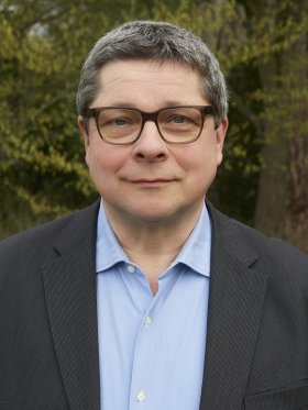 Ein Mann mittleren Alters steht im Freien. Er trägt ein hellblaues Hemd und ein dunkelgraues Sakko. Hinter ihm sind grüne Bäume und Büsche zu sehen, die eine natürliche Kulisse bilden
