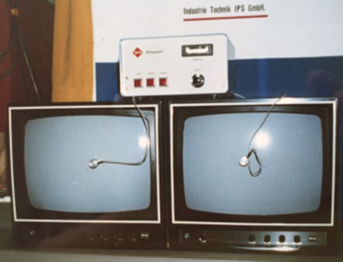Zwei Röhrenfernseher mit blauen Bildschirmen zeigen jeweils ein Kabel mit rundem Metallgegenstand. Darüber befindet sich ein weißes Gerät mit Schaltern und Drehknopf