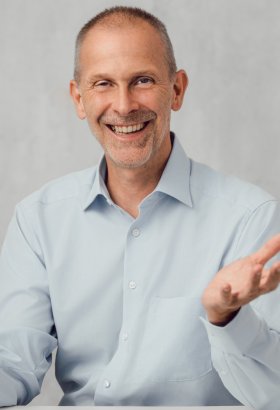 Etwas älterer Herr mit sehr kurzen Haaren und offenem hellblauem Hemd an einem Tisch sitzend