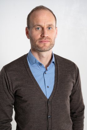 Porträt einer Person mit kurzen Haaren und leichtem Bart, hellblauem gemusterten Hemd und brauner Strickjacke vor neutralem, weißem Hintergrund