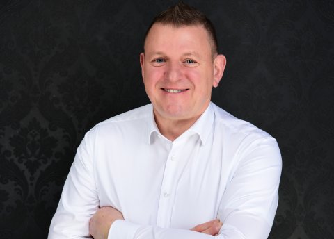 Person in weißem Hemd mit verschränkten Armen vor dunklem, gemustertem Hintergrund