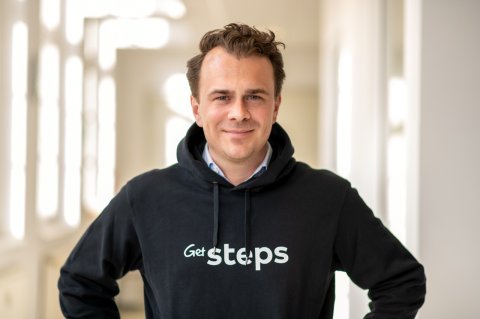 Person trägt einen schwarzen Hoodie mit dem GetSteps Logo in einem hellen Flur