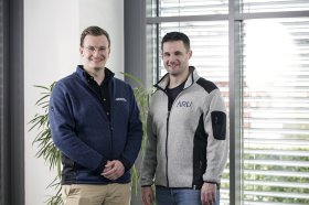 Zwei Personen stehen nebeneinander in einem Bürobereich und tragen Arbeitsjacken mit Firmenlogo vor einem Fenster mit Jalousien