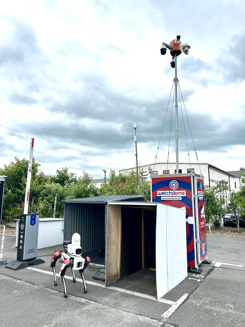 Roboterhund vor Kleingarage, im Hintergrund ein Mast mit Kameras