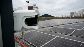 Security camera mounted at a solar farm, overlooking rows of solar panels with visible cable routes and buildings in the background