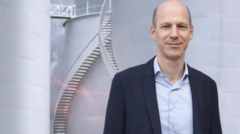 Porträt einer Person vor großen Industrietanks mit Außentreppe; im Vordergrund eine grafische Visualisierung von Sensordaten.